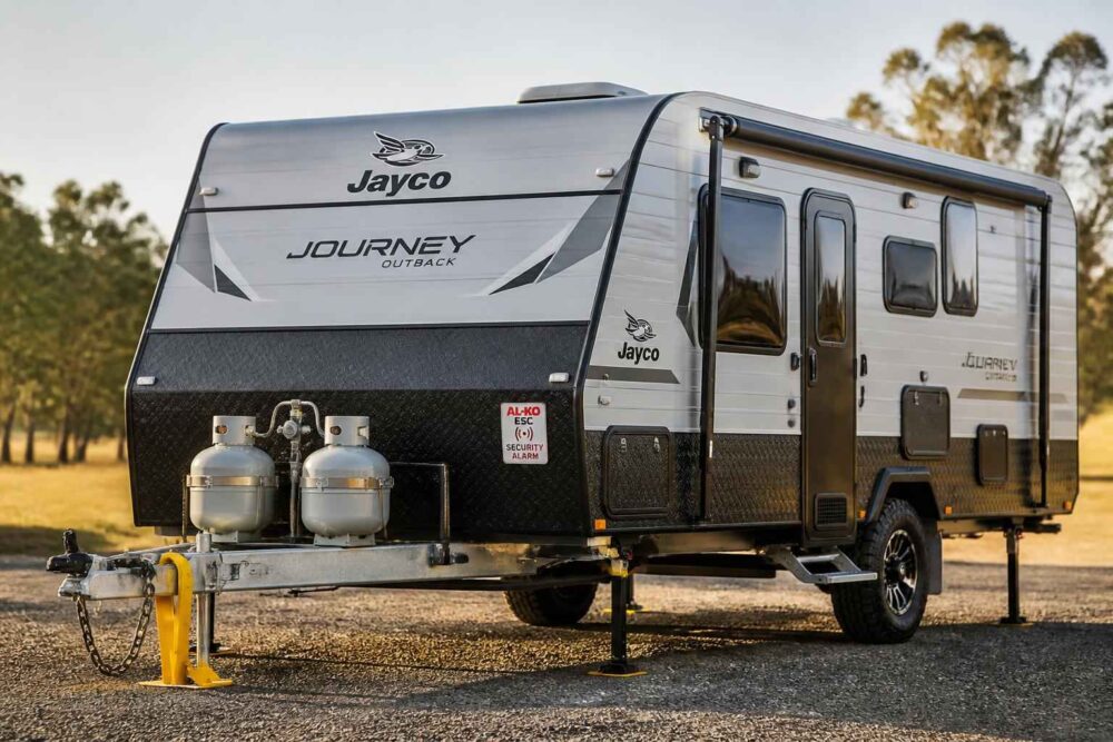 Srebrna przyczepa kempingowa Jayco Journey zaparkowana na zewnątrz pod drzewami